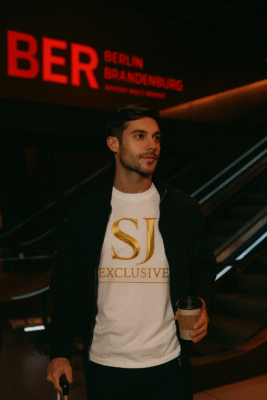 Homme chic à l’aéroport de Berlin portant le t-shirt blanc SJ EXCLUSIVE avec logo doré, style premium en voyage.